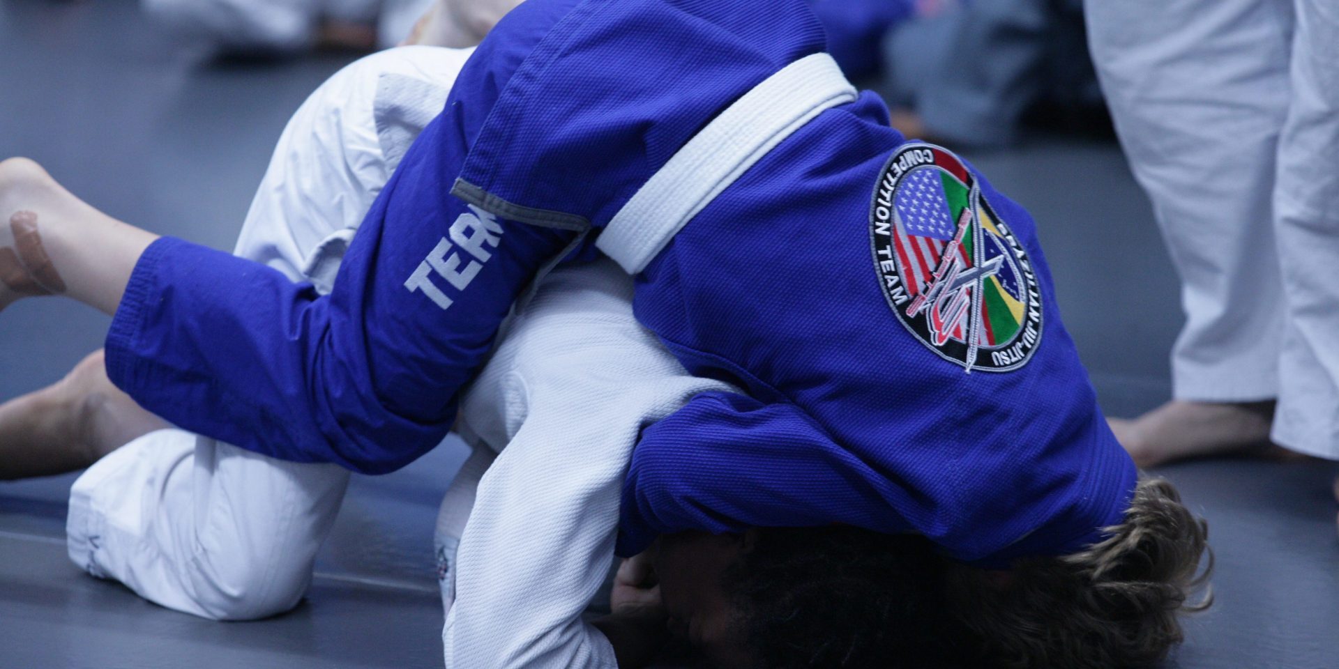 Two individuals grappling in Brazilian Jiu-Jitsu training, wearing blue and white gis.